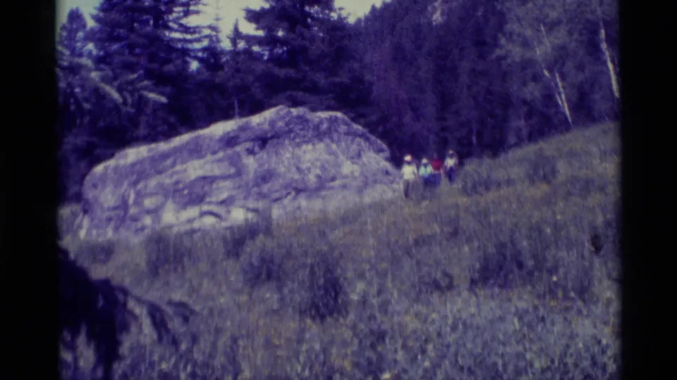 Wide 1975 8mm view of three hikers crossing grass near a large boulder, with pine forest behind in Agate Springs, Montana.