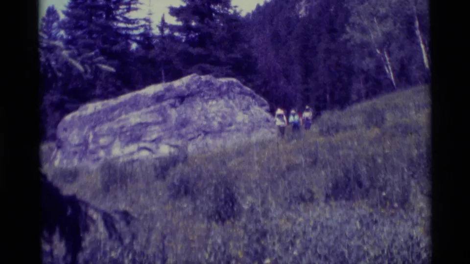 Three hikers continue across a grassy Montana hillside beside a large rock, seen in soft, purple-tinted 8mm home-movie footage.