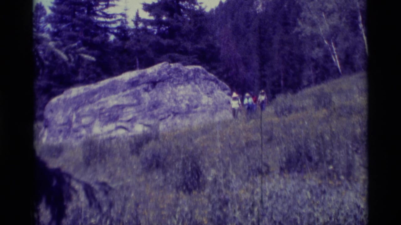 1975 Agate Springs, Montana home-movie frame showing hikers approaching along a grassy trail beside a large rock formation.