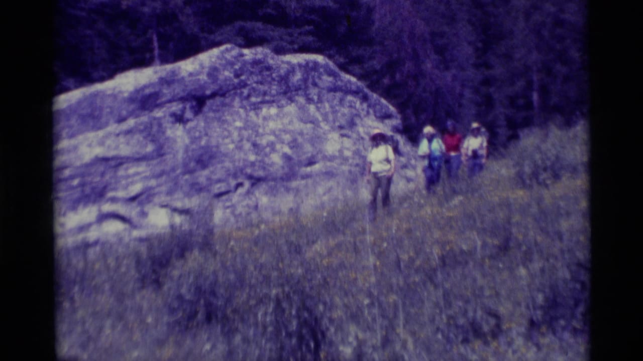 Backpackers walk beside a large rock formation on a grassy trail.