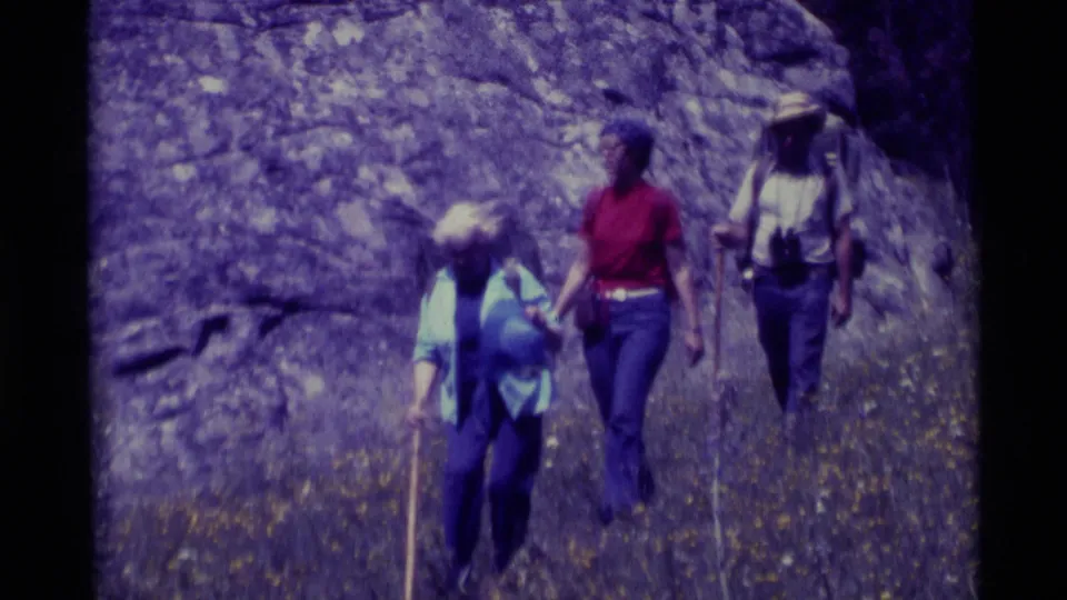 1975 Agate Springs, Montana frame with hikers in casual 1970s clothing moving uphill near a textured rock face.