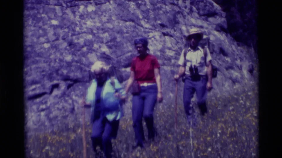 Three hikers in 1975 Agate Springs, Montana move across a rocky slope with sticks, a pack, and binoculars visible.