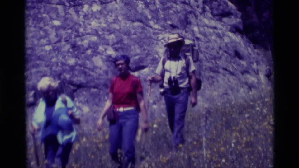 The hiking group continues through rocky Agate Springs terrain, with a woman in red leading and a man in a straw hat behind.
