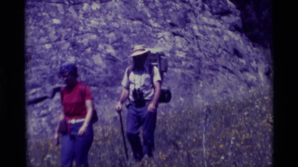A woman in red and a man with a straw hat, pack, binoculars, and walking stick pass near the camera in Montana hiking footage.