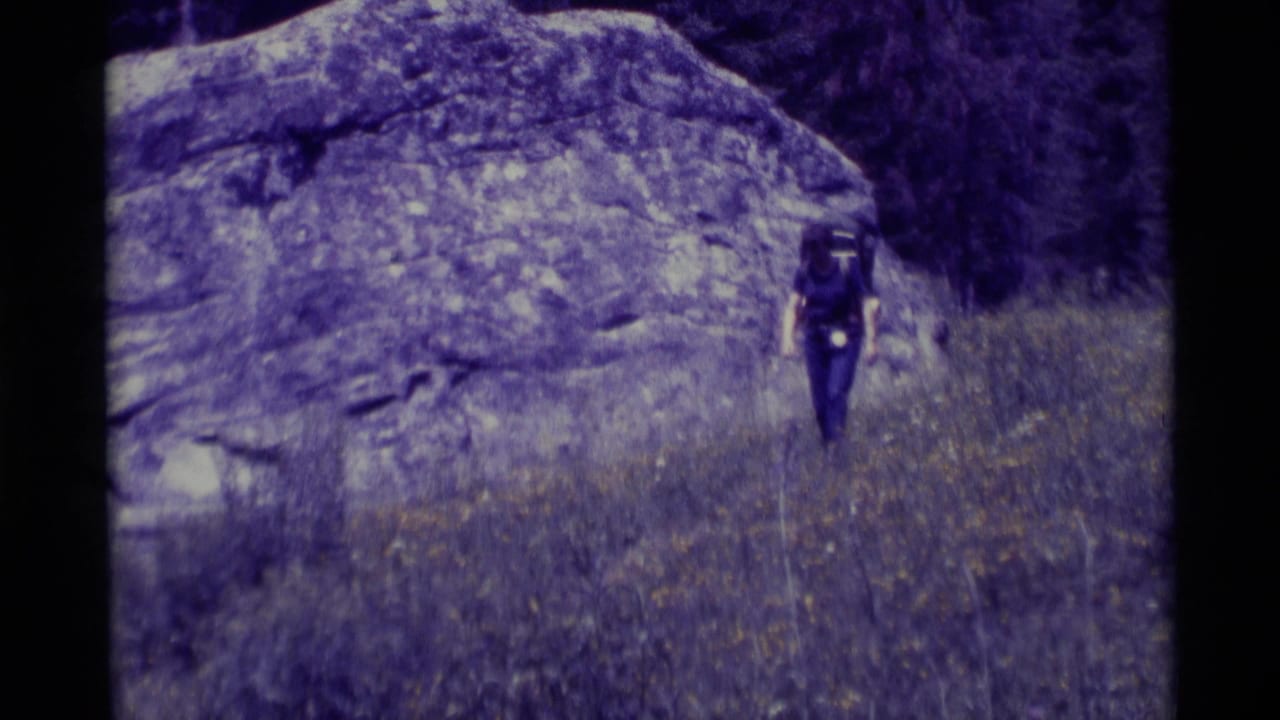 Woman with a large backpack walking through grass below a rock face.