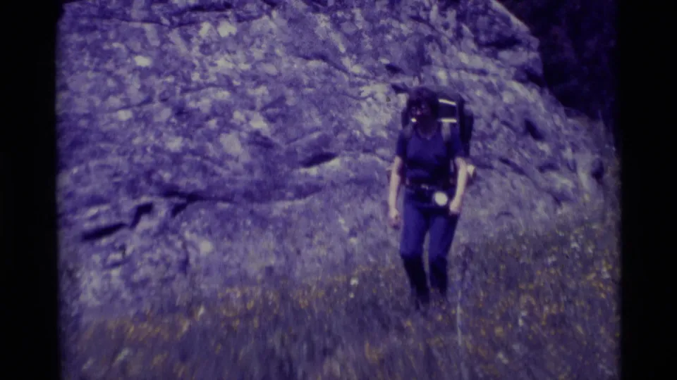 A woman carrying a large backpack walks toward the camera in purple-shifted 1975 Montana home-movie footage.
