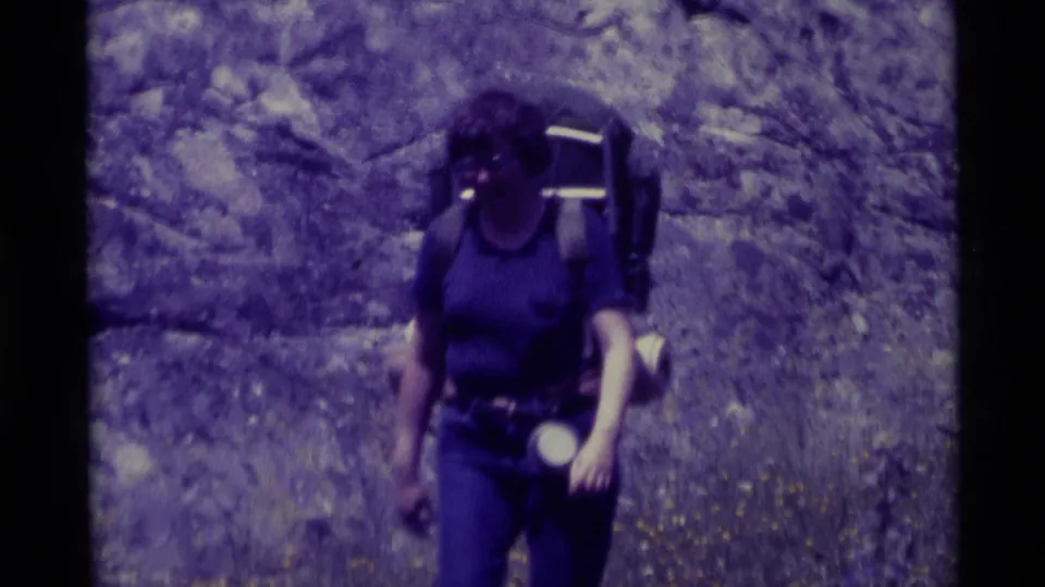 Close 8mm frame of the woman backpacker, with her pack, belt gear, and flashlight visible as she passes the camera.
