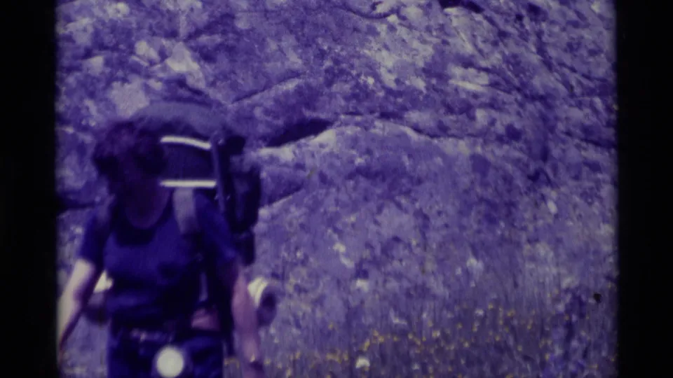 The woman hiker passes near the camera, partly cropped, with her backpack and flashlight visible against the rocky Montana setting.