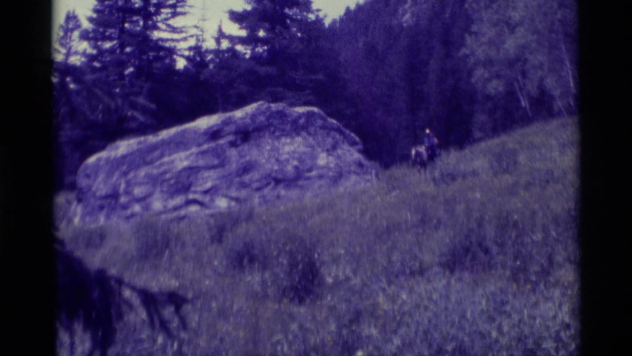 1975 8mm home-movie frame from Agate Springs, Montana, showing two distant horseback riders near a large boulder in a meadow.