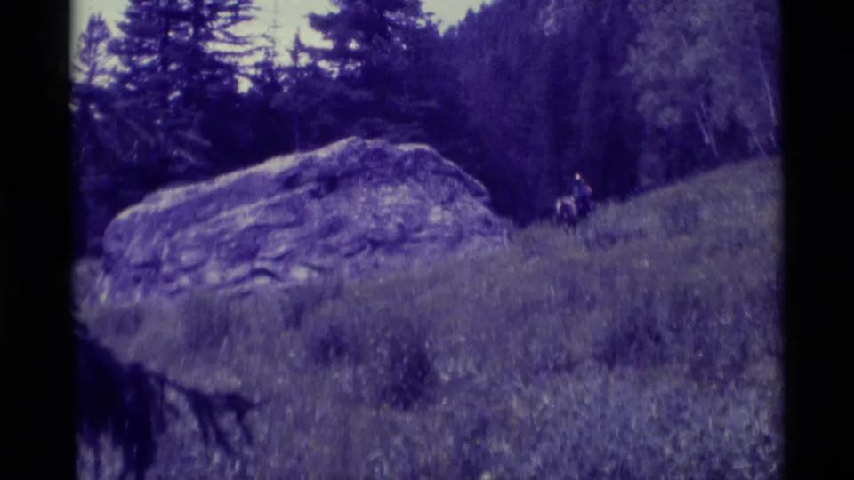 1975 8mm home-movie frame from Agate Springs, Montana, showing two distant horseback riders near a large boulder in a meadow.