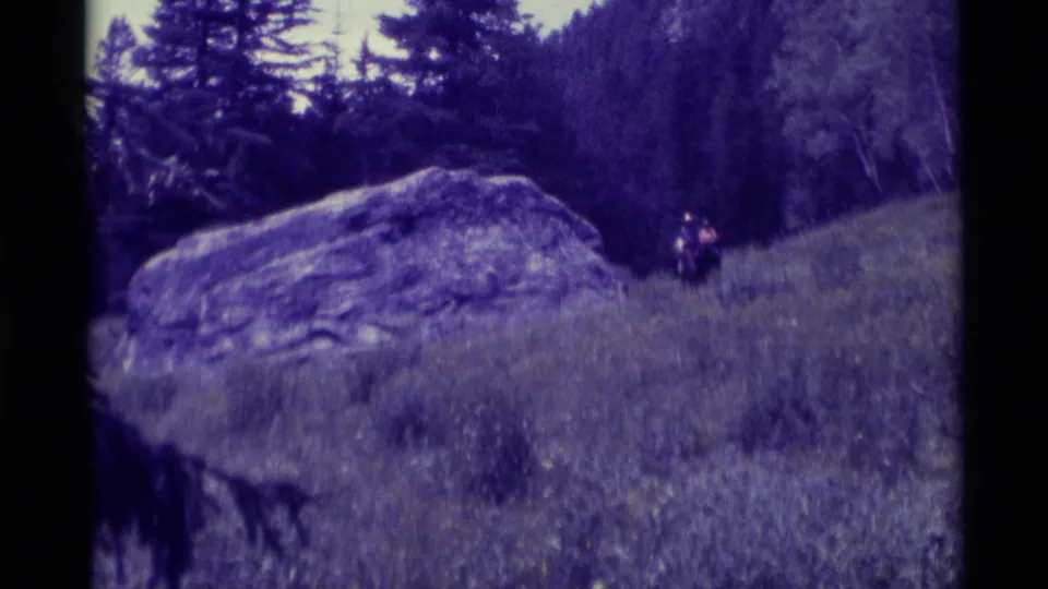 Distant horseback riders cross a meadow near a large boulder in the supplied 1975 Agate Springs, Montana home-movie footage.