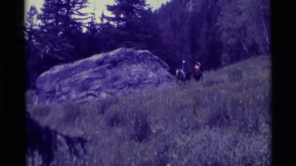 Final frame of the 1975 Montana 8mm clip, with two distant riders moving through grass beside a large boulder and wooded backdrop.