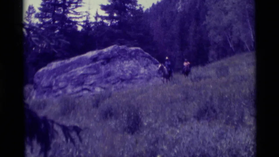 1975 8mm Montana home-movie frame showing two distant riders on horseback near a large boulder and trees.