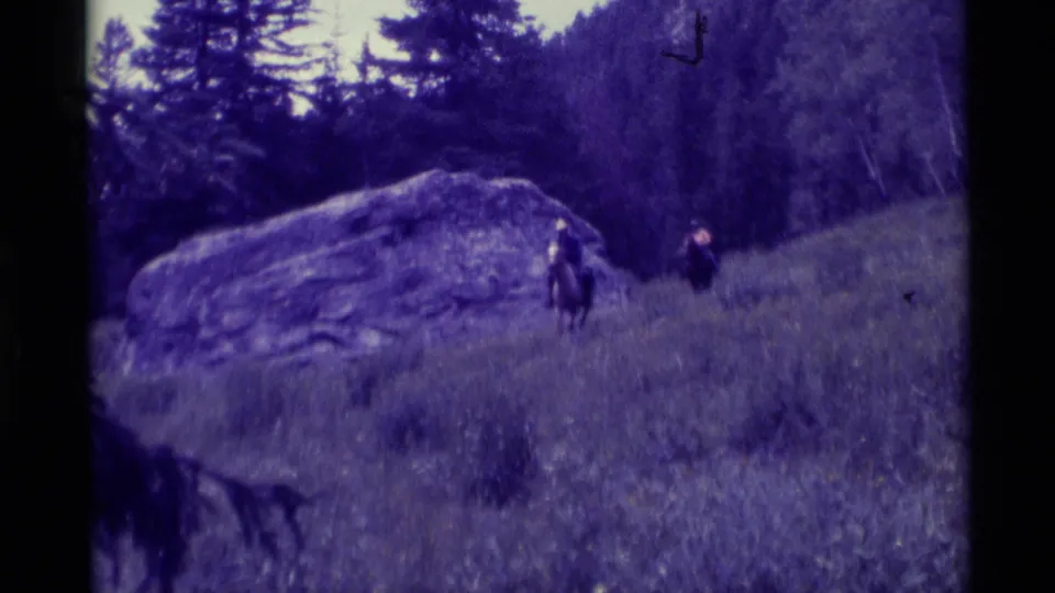 Two riders on horseback move across a grassy Agate Springs hillside in soft purple-tinted 8mm footage.