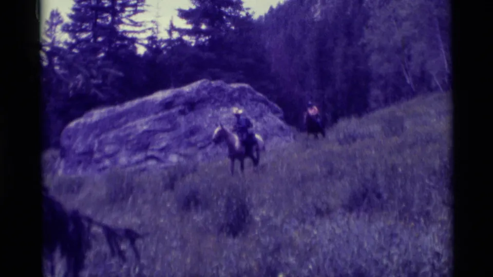 A light-hatted rider on horseback passes near a large boulder, with a second rider farther up the grassy slope.