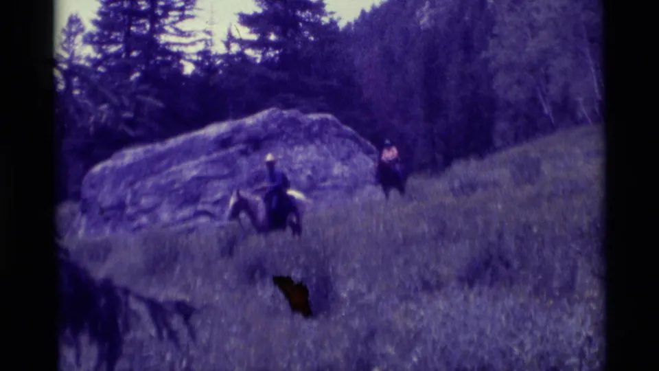 End frame of two horseback riders crossing a Montana hillside, framed by a boulder, trees, and 8mm scan borders.