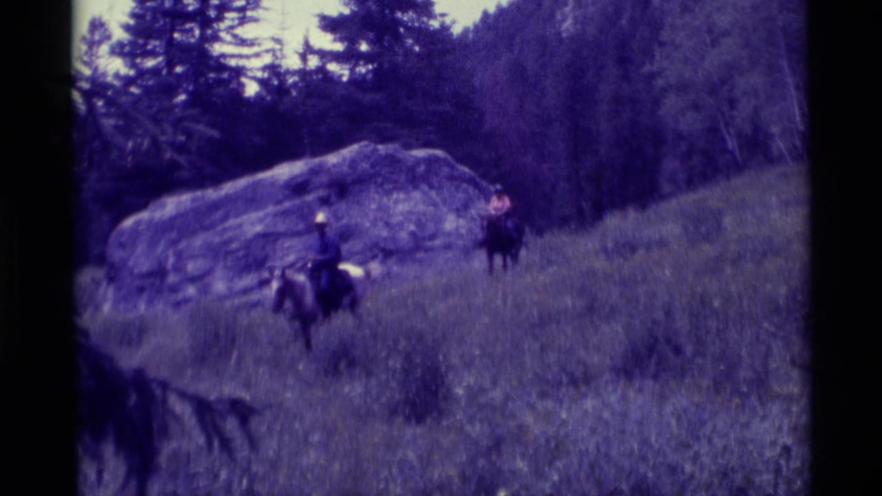 1975 8mm frame from Agate Springs, Montana, showing three men riding horses on a grassy slope beside a large rock and evergreens.