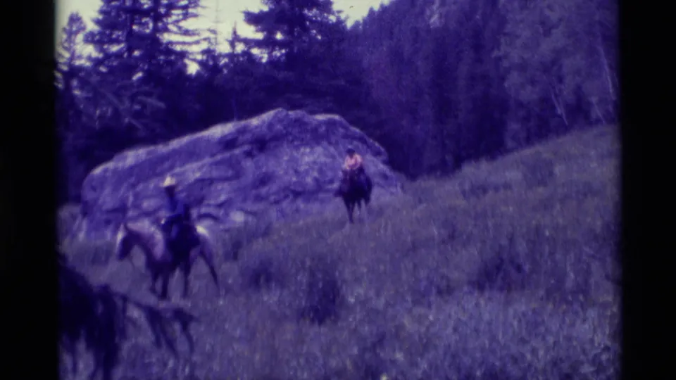 A 1975 Montana home-movie frame shows riders on horseback crossing tall grass below a boulder and wooded slope.