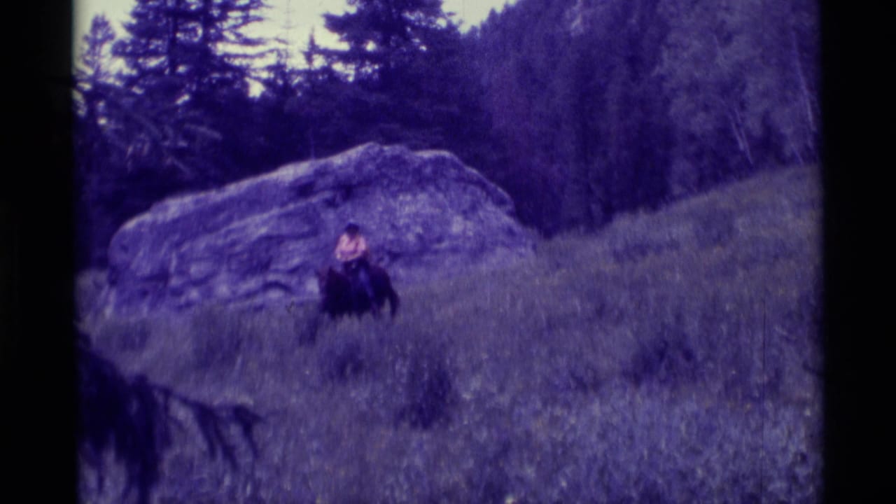 1975 8mm home-movie frame from Agate Springs, Montana, showing a rider on horseback crossing a grassy mountain slope near a boulder.