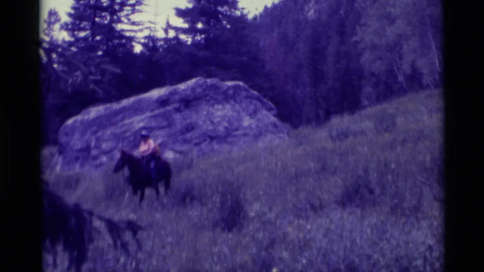 The rider and horse appear mid-frame against a grassy Montana hillside, with a large boulder and evergreen trees behind them.