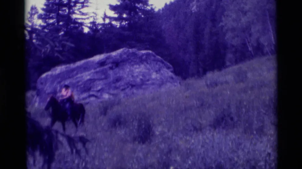 In this 1975 Montana home-movie frame, the rider on horseback nears the left frame edge below the boulder and trees.