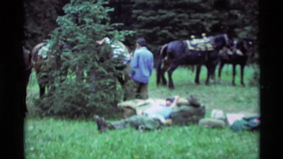 A 1975 Montana home-movie frame shows one person resting near packs as horses and another person stand deeper in the forest clearing.
