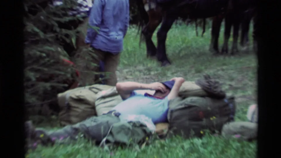 Closer 1975 Montana frame of a man in a blue shirt and dark hat resting on camping gear near saddled horses in grass.