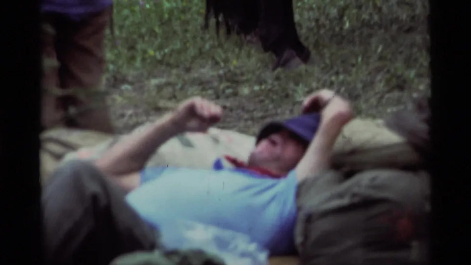 1975 Montana home-movie close frame of a man reclining on packs, with nearby horse legs and grassy ground visible.