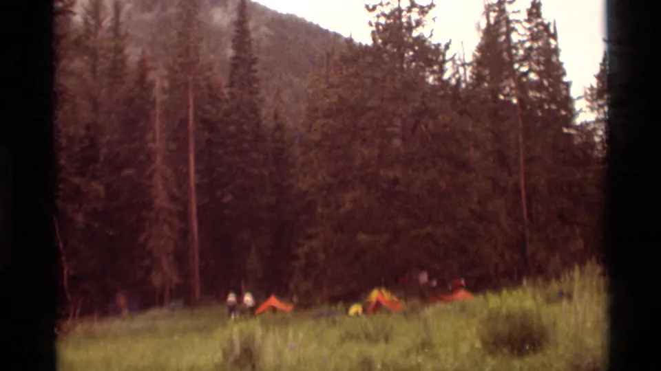 Orange tents and distant campers appear in a grassy Agate Springs meadow below tall pines and a forested slope.