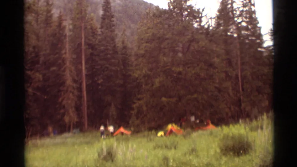 A wide 1975 Montana campsite view shows orange tents and small figures across a meadow bordered by tall forest.