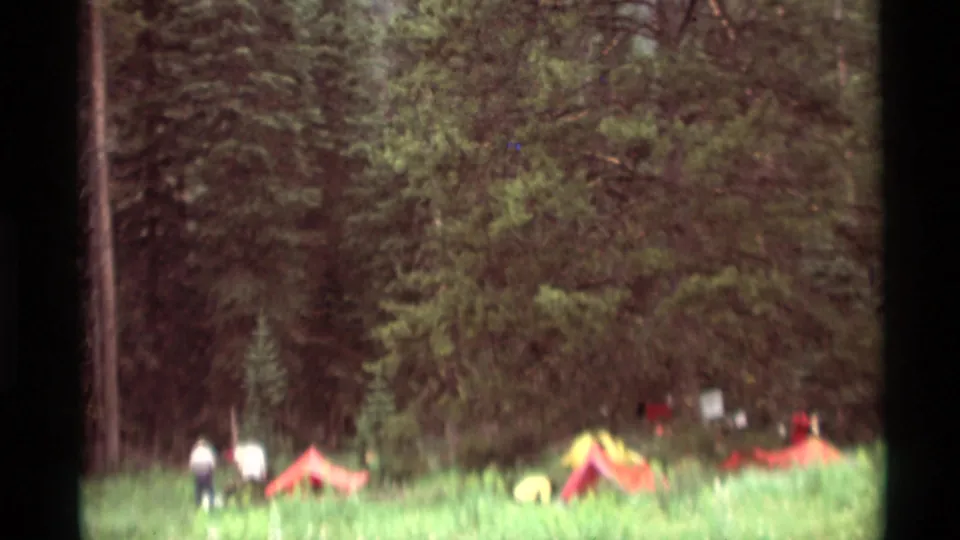 Soft 1970s 8mm frame showing a Montana campsite with small tents and distant figures near the woods.
