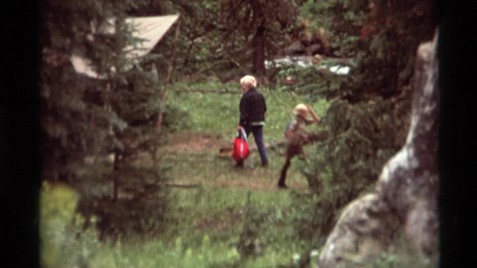 At the edge of the Montana campsite, two figures walk along a path while one carries a red bag.