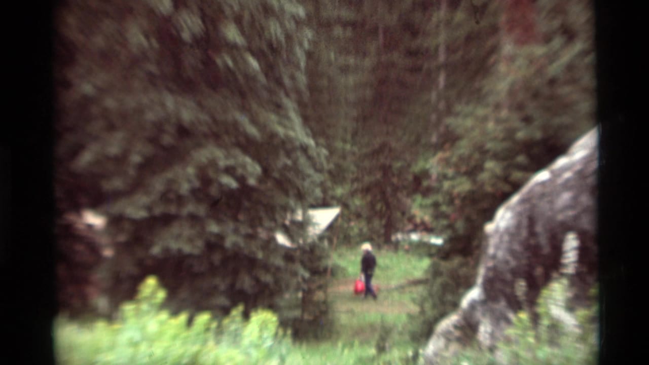 1975 8mm home-movie frame from Agate Springs, Montana, showing a distant walker among pines and grass with a red bag.