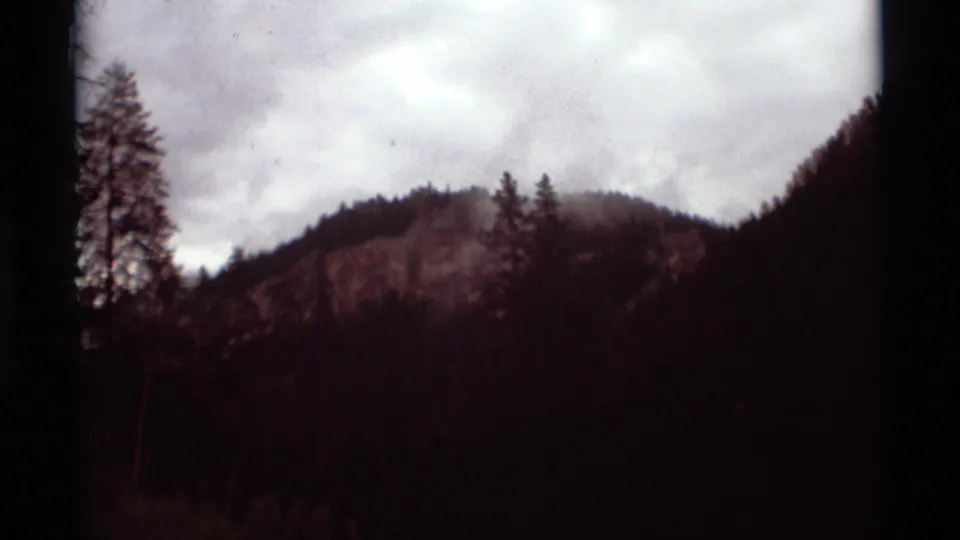 1975 8mm mountain landscape near Agate Springs, Montana, with cloudy sky, dark trees, and a pale rock face.