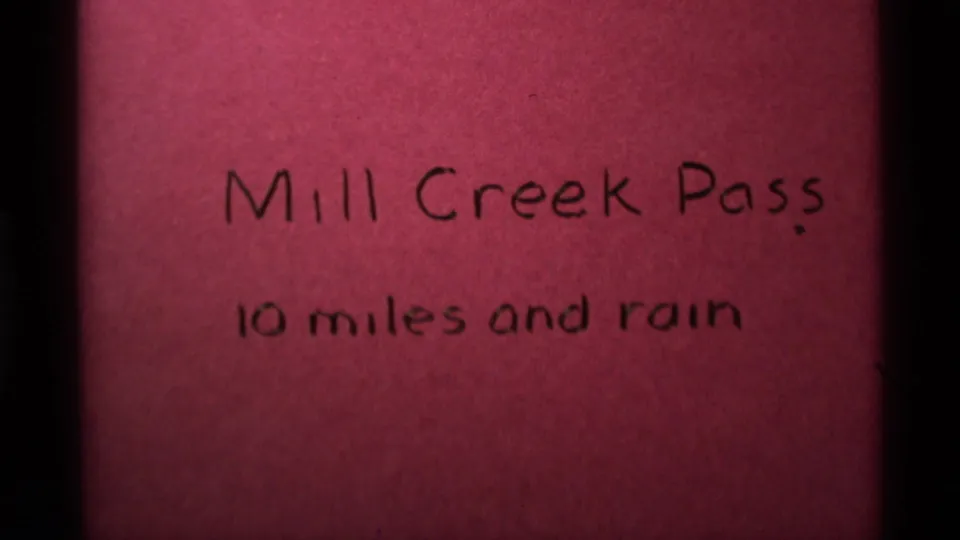 Closer 8mm frame of the handwritten Mill Creek Pass title card, with the rain note centered below.