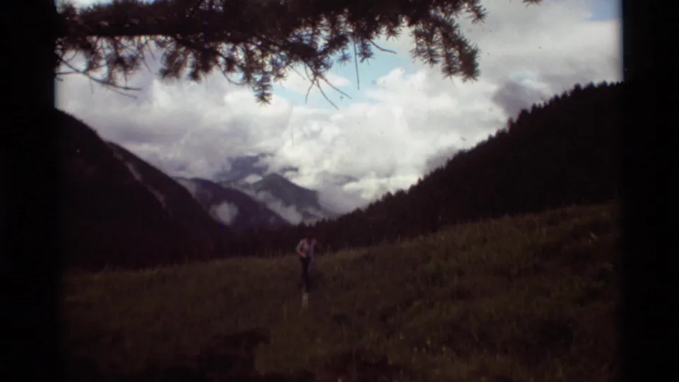 1975 Montana frame showing a lone hiker in an open valley with pine branches overhead and cloudy mountains beyond.