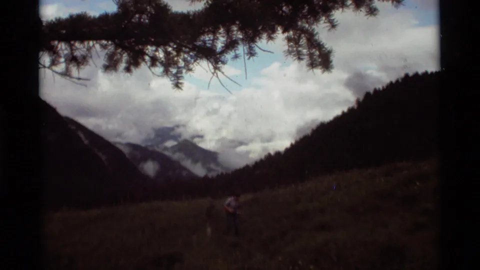 Pine branches frame a 1975 Montana mountain valley view with cloud-covered peaks and a small distant person in the meadow.