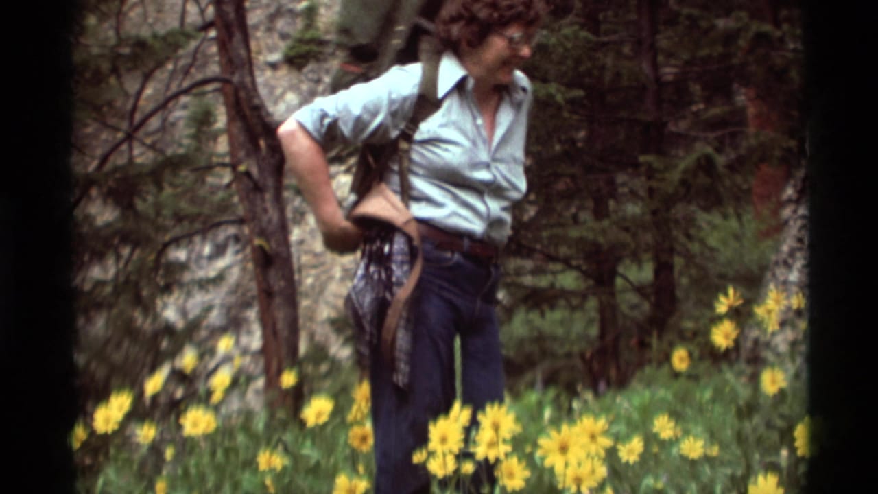 In Agate Springs, Montana, 1975, a woman hiker stands in a flowered mountain meadow adjusting her backpack.
