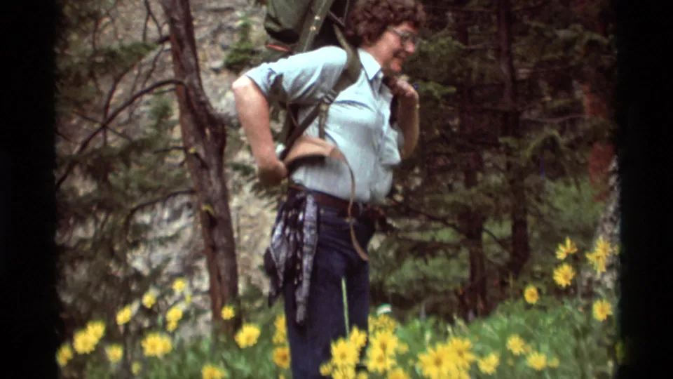 A woman hiker in 1975 Montana adjusts her backpack straps in a meadow bordered by pine trees and rocky ground.