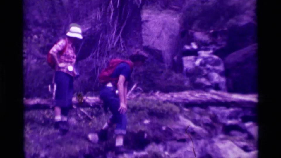 In the Agate Springs 1975 home movie, one figure bends toward the stream bank while another watches near a fallen log.