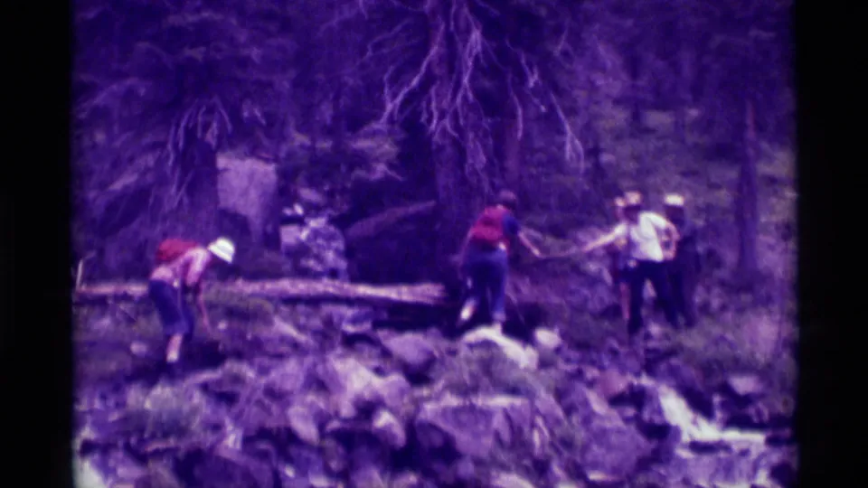 Purple-shifted 8mm frame showing hikers navigating rocks and a log on a wooded Montana trail during a 1975 family outing.