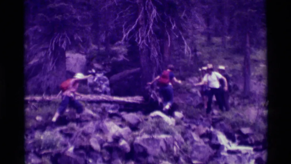 Hikers in a purple-tinted 1975 8mm frame move across a rocky wooded trail, with a fallen log and forest growth behind them.