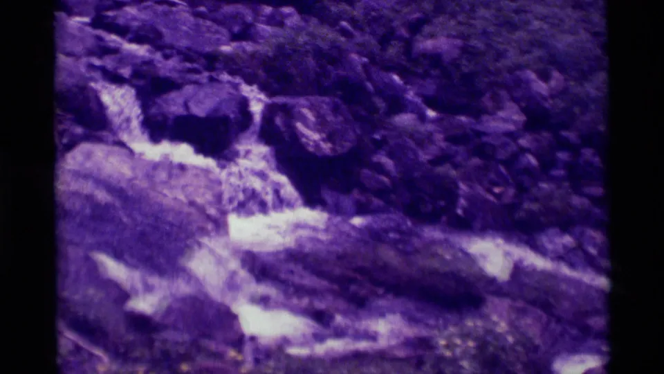 1975 8mm home-movie frame from Agate Springs, Montana, showing white water moving through dark rocks with purple fading.