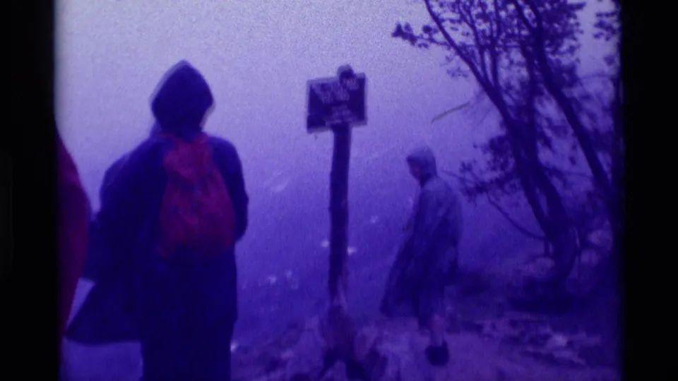 Hikers with rain gear and backpacks gather by a Mill Creek Pass elevation sign in foggy 1975 Montana 8mm home-movie footage.