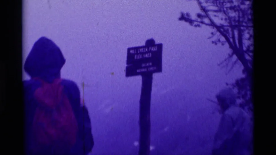 Hooded hiker in rain gear stands before the Mill Creek Pass Elev. 9403 sign during a foggy 1975 Montana mountain stop.