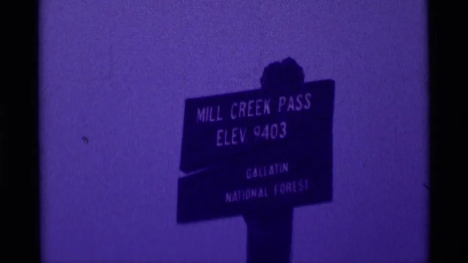 Close 8mm view of the Mill Creek Pass Elev. 9403 Gallatin National Forest sign against a purple foggy sky.