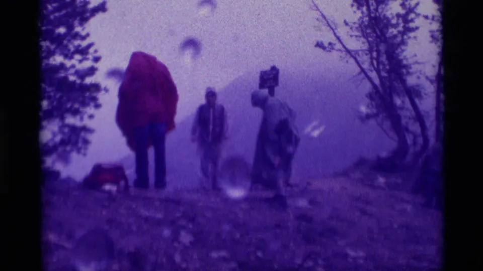 1975 Montana home-movie frame showing three visitors in rainwear on a misty mountain trail near Mill Creek Pass.