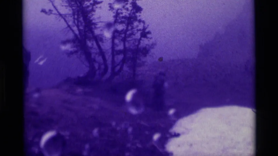 1975 8mm frame of the Mill Creek Pass area, showing a misty trail, trees, snow patches, and water-marked lens texture.