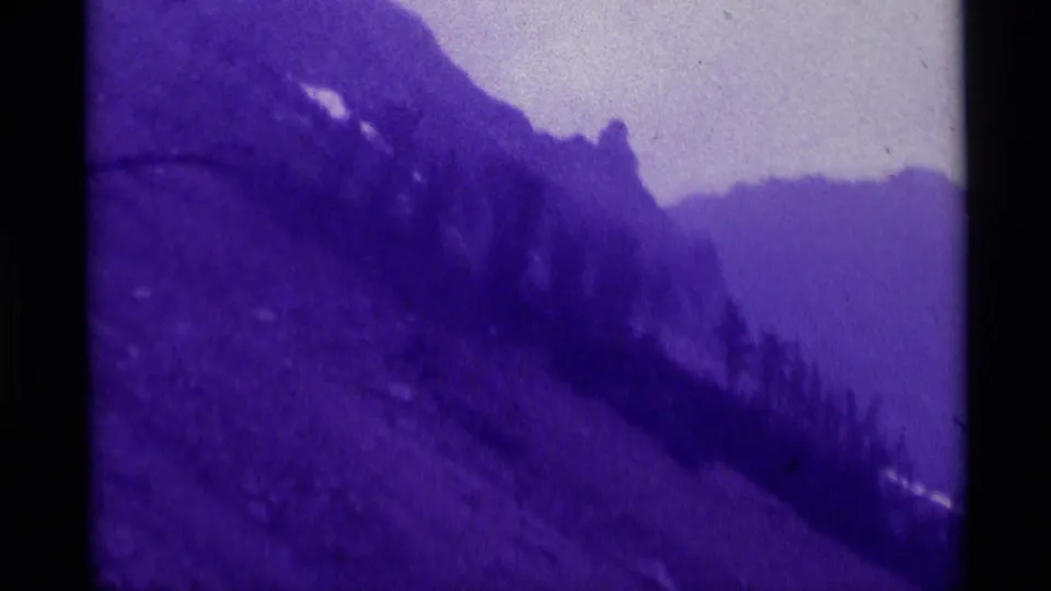 Purple-cast 1975 Montana 8mm frame with a distant person on a sloped mountainside beneath pale sky and hazy ridges.