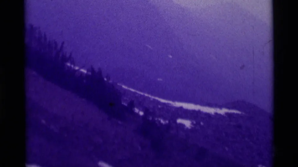 1975 8mm frame of a Montana mountain landscape, with snow patches, dark trees, and a faint possible road or trail along the lower slope.